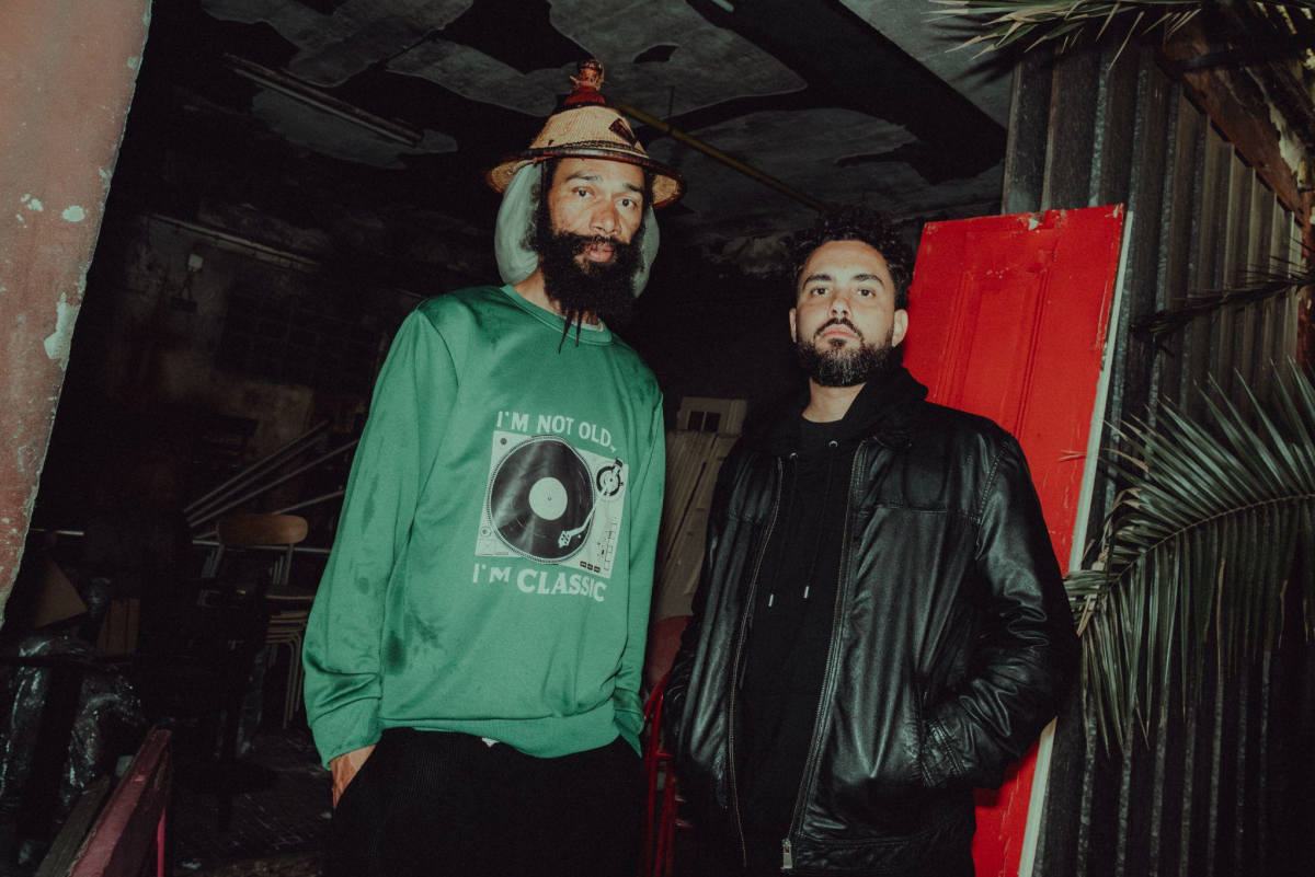 A photo of two men looking straight into the camera. They both have dark beards. The man on the left is wearing a hat and a green sweater. The man on the right is wearing a black hoodie and a black leather jacket.